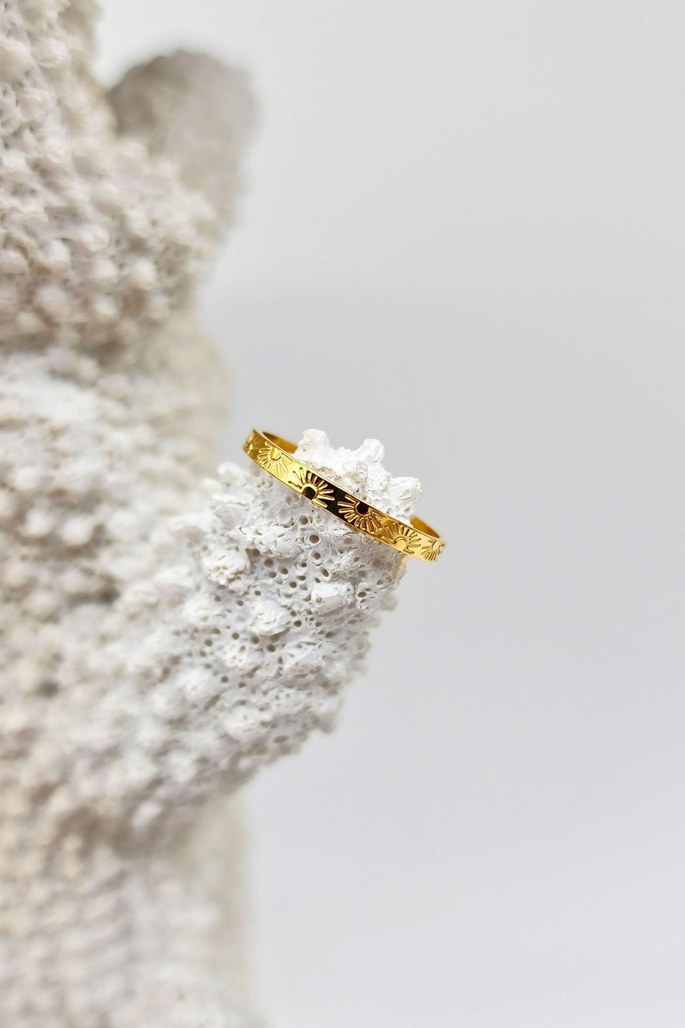Gold band ring with sun and ray designs, beautifully placed atop a white coral piece.