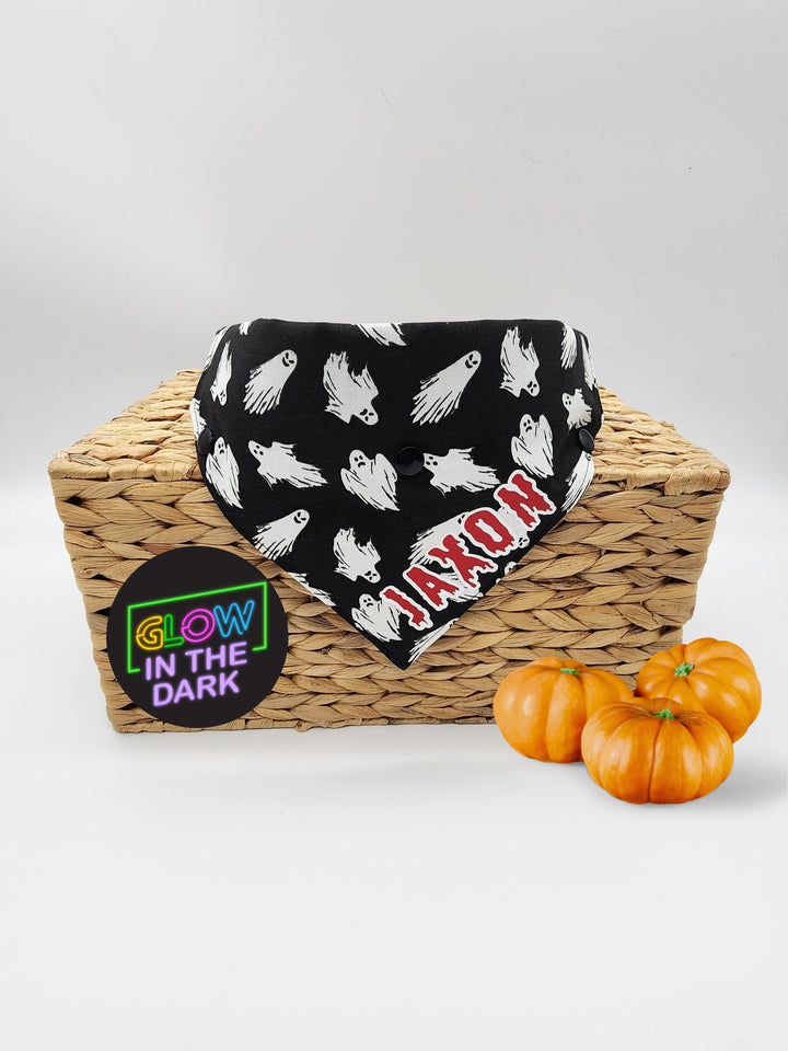 Displayed atop of a wooden box is black halloween dog bandana with glow in the dark print of floating ghosts a red and white graphic that reads the name Jaxon.