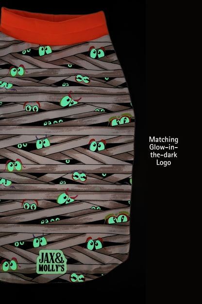 Glow-in-the-dark dog pajama with eyes peeking out from behind woven strands and 'Jax & Molly's' branding, set against a dark background.