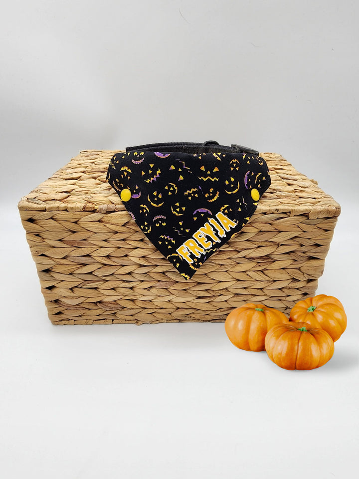 Displayed atop of a wooden box is a black halloween dog bandana featuring lit up pumpkin faces and a yellow graphic that reads the name Freyja.