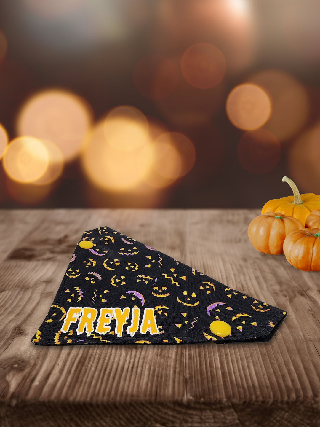 Displayed on a wooden table is the side profile of black halloween dog bandana featuring lit up pumpkin faces and a yellow graphic that reads the name Freyja.