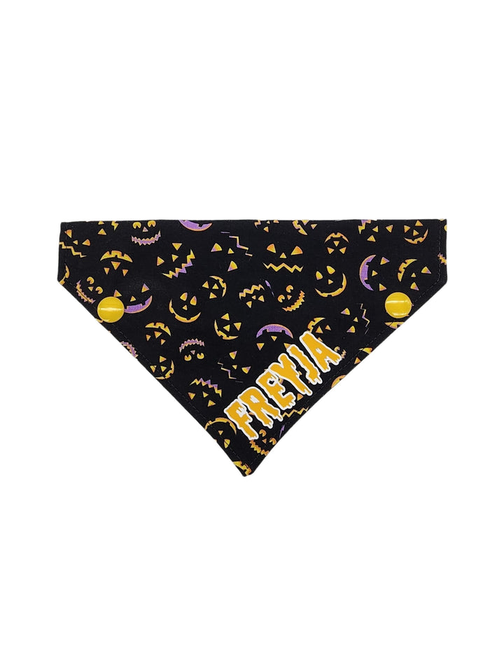 Front profile of black halloween dog bandana featuring lit up pumpkin faces and a yellow graphic that reads the name Freyja on a white background.