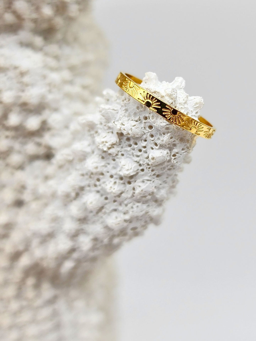 Gold band ring with sun and ray designs, beautifully placed atop a white coral piece.