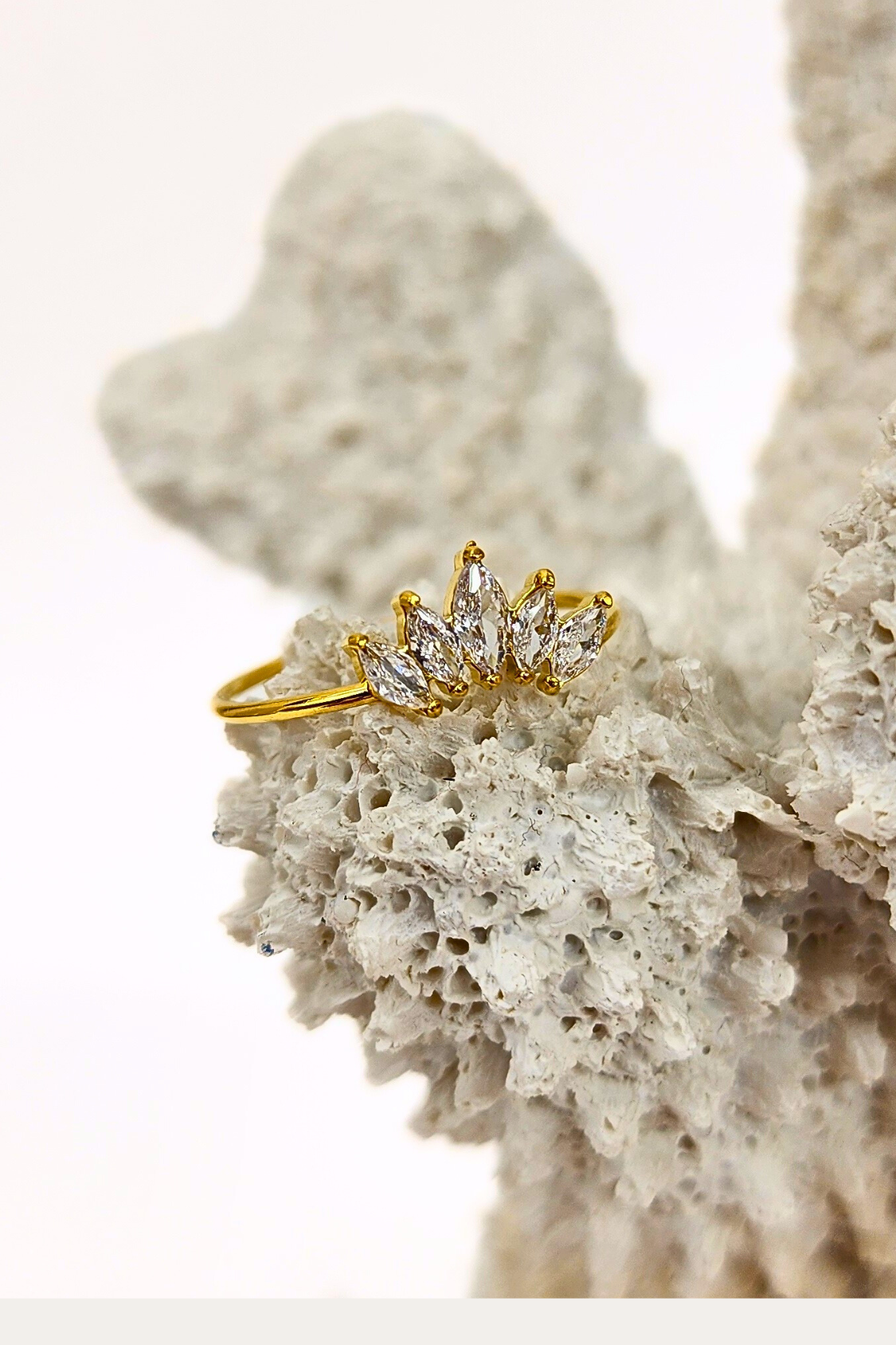 Regal Radiance ring featuring a crown-like arrangement of 5 clear gemstones on a thin 18k PVD gold plated band, elegantly placed on white coral for a stylish display.