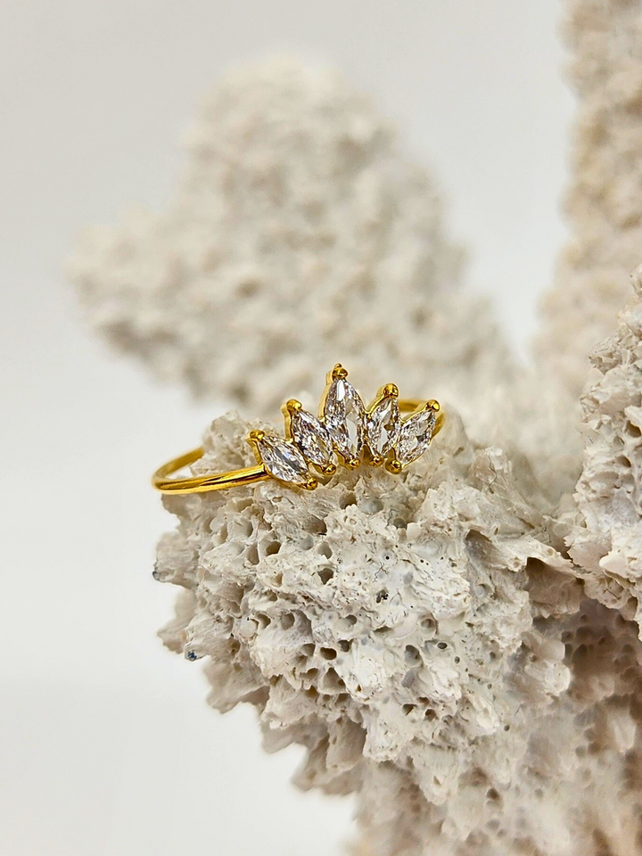 Regal Radiance ring featuring a crown-like arrangement of 5 clear gemstones on a thin 18k PVD gold plated band, elegantly placed on white coral for a stylish display.