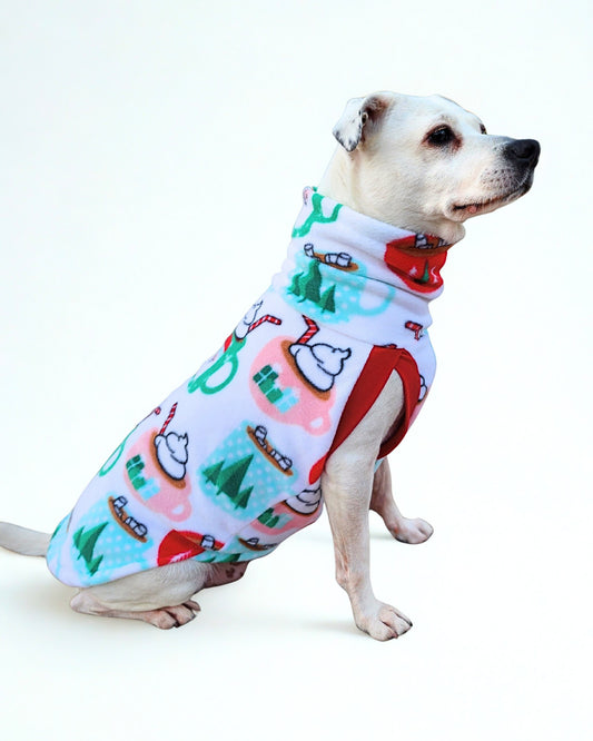 Side profile of a white dog wearing a cute sweater with a print of cocoa mugs adorned with Santa faces and pine trees on white.