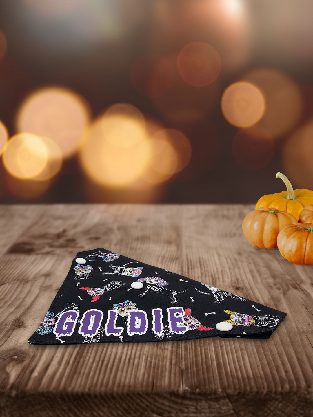 Displayed on a wooden table is the side profile of a black halloween dog bandana featuring a sugar skull dog skeleton pattern and a purple graphic that reads the name Goldie.